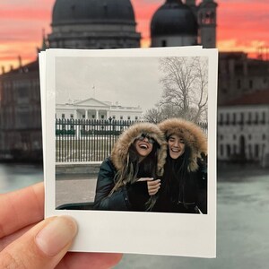 May include: Two women wearing winter coats with fur hoods are smiling and looking at the camera. They are standing in front of a white building with a black fence. The building has a white roof and a flagpole with a flag on top.