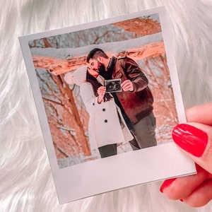 May include: A couple in a snowy forest holding an ultrasound photo. The woman is wearing a white coat and a white knit hat. The man is wearing a brown leather jacket. The photo is in a white frame.