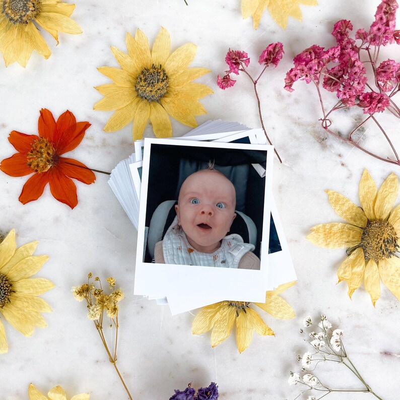 May include: A close-up photo of a baby boy with blonde hair and blue eyes. He is wearing a white shirt with a blue and white pattern. The photo is printed on a white polaroid-style photo card.