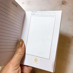 Op de afbeelding: Open notitieboek met gelinieerde pagina's en het woord "NOTES". Er is een sectie voor "PLACE:" en "DATE:" en een leeg vak. Het notitieboek heeft een patroonrand en een klein logo. De pagina's zijn wit en de kaft is lichtbeige.