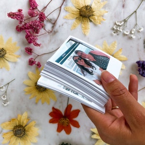 May include: A stack of white photo prints with a photo of two people smiling at the camera on top. The photo shows two women, one with dark hair and sunglasses, and the other with gray hair. The photo is held in a hand.