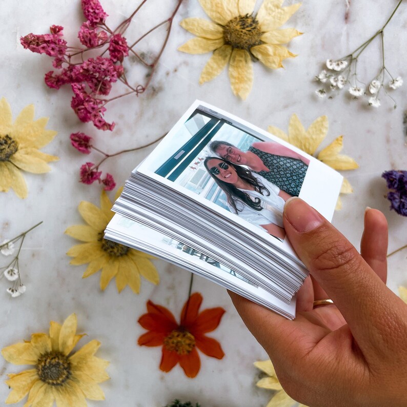 May include: A stack of white photo prints with a photo of two people smiling at the camera on top. The photo shows two women, one with dark hair and sunglasses, and the other with gray hair. The photo is held in a hand.