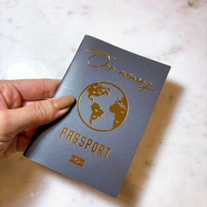 Op de afbeelding: Een grijze paspoort met gouden folie letters en een wereldkaart ontwerp. De cover leest "Our memory" in cursief en "PASSPORT" in hoofdletters. Een klein rechthoekig embleem staat onder het woord "PASSPORT".