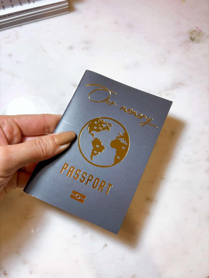 Op de afbeelding: Een grijze paspoort met gouden folie letters en een wereldkaart ontwerp. De cover leest "Our memory" in cursief en "PASSPORT" in hoofdletters. Een klein rechthoekig embleem staat onder het woord "PASSPORT".
