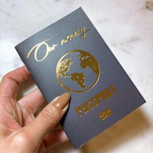 Op de afbeelding: Een donkergrijze paspoort met gouden letters en een wereldkaartontwerp. De tekst "Our memory" staat bovenaan en "PASSPORT" is onder de kaart gedrukt. Onderaan staat een klein gouden embleem.