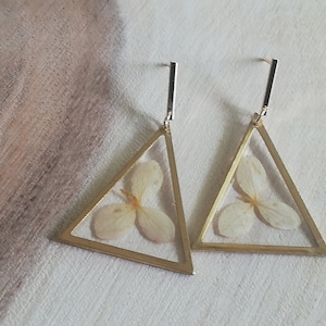 Può includere: Un paio di orecchini triangolari dorati con fiori bianchi secchi pressati all'interno della resina.