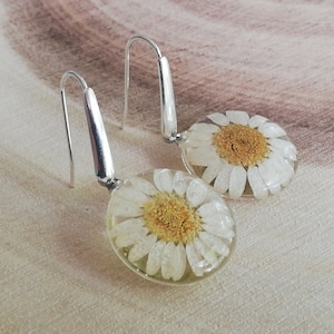 May include: A pair of silver dangle earrings with a white daisy pressed inside a clear resin oval.