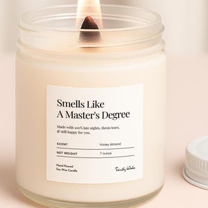 May include: A lit, honey almond-scented candle in a clear glass jar with a white label that reads "Smells Like A Master's Degree." The jar is next to a white lid. The candle is 198 grams.