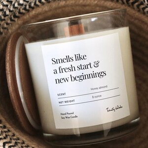 May include: A clear glass candle jar with a wooden lid. The candle is white and has a label that reads "Smells like a fresh start & new beginnings". The label also includes the scent, "Honey almond", and the net weight, "8 ounce". The candle is sitting in a brown and black woven basket.