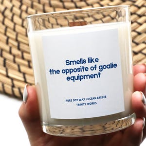 May include: A white candle in a clear glass jar with a white label that reads "Smells like the opposite of goalie equipment". The label also includes the text "Pure Soy Wax | Ocean Breeze" and "Trinity Works".