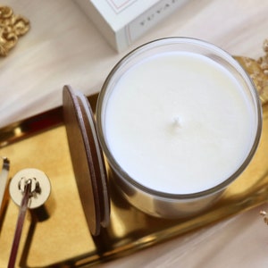 May include: A white candle in a clear glass jar sits on a gold tray with a wooden lid. The tray is on a white surface with dried flowers.