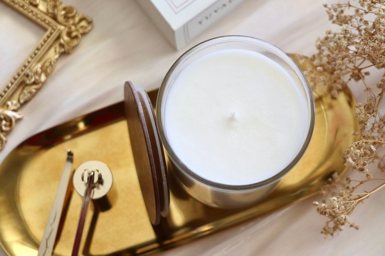 May include: A white candle in a clear glass jar sits on a gold tray with a wooden lid. The tray is on a white surface with dried flowers.