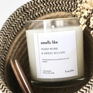 May include: A clear glass candle with a white label that reads "smells like HARD WORK & SWEET SUCCESS Congratulations! I am so excited for your next chapter! Honey Almond Soy Wax Candle Trinity Wodo". The candle is sitting in a woven basket with a black and white pattern.
