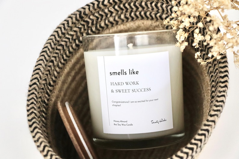 May include: A clear glass candle with a white label that reads "smells like HARD WORK & SWEET SUCCESS Congratulations! I am so excited for your next chapter! Honey Almond Soy Wax Candle Trinity Wodo". The candle is sitting in a woven basket with a black and white pattern.