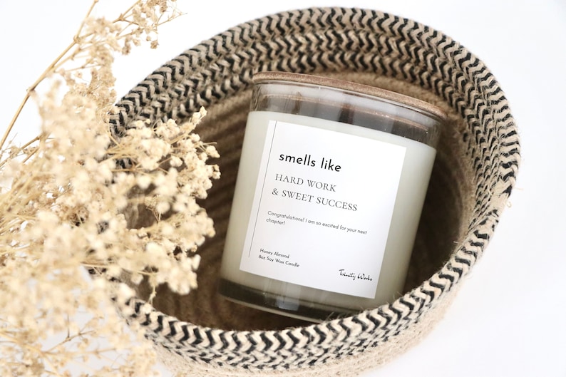 May include: A white soy wax candle in a clear glass jar with a black and white label that reads "smells like HARD WORK & SWEET SUCCESS Congratulations! I am so excited for your next chapter! Honey Almond Soy Wax Candle Trinity & Wade". The candle is sitting in a woven basket with black and white stripes.