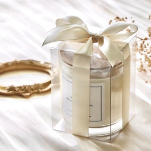 May include: A clear glass candle jar with a white candle inside. The jar is tied with a cream-colored satin ribbon and bow. The candle has a white label with the text "& Mademoiselle" printed on it.