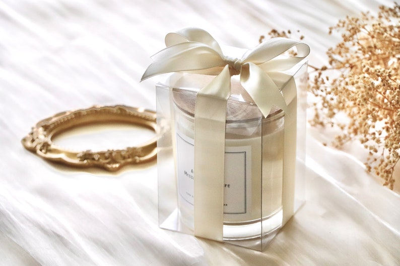 May include: A clear glass candle jar with a white candle inside. The jar is tied with a cream-colored satin ribbon and bow. The candle has a white label with the text "& Mademoiselle" printed on it.