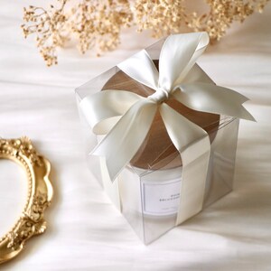 May include: A white candle in a clear glass jar with a wooden lid, wrapped in a clear box with a white satin ribbon bow.