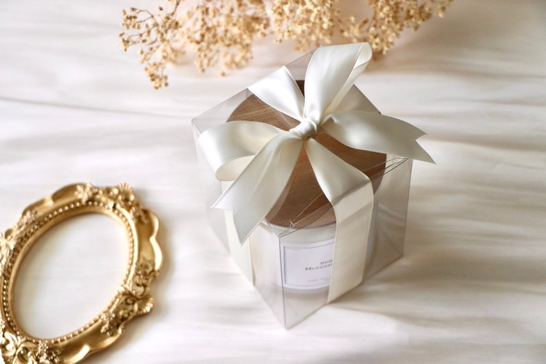 May include: A white candle in a clear glass jar with a wooden lid, wrapped in a clear box with a white satin ribbon bow.