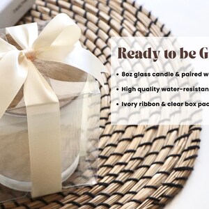 May include: A clear box with a white ribbon tied around it, containing a glass candle with a wooden lid. The candle is on a woven mat. The text "Ready to be Gifted!" is written in black on a white background. The text below lists the features of the candle: "8oz glass candle & paired with wood lid", "High quality water-resistant label", and "Ivory ribbon & clear box packaging".