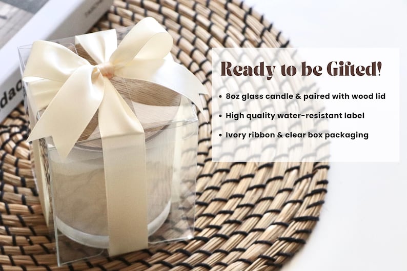 May include: A clear box with a white ribbon tied around it, containing a glass candle with a wooden lid. The candle is on a woven mat. The text "Ready to be Gifted!" is written in black on a white background. The text below lists the features of the candle: "8oz glass candle & paired with wood lid", "High quality water-resistant label", and "Ivory ribbon & clear box packaging".