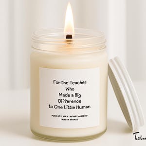 May include: A burning candle in a glass jar with a white label. The label reads "For the Teacher Who Made a Big Difference to One Little Human." The lid is off to the side. The candle is honey almond scented.