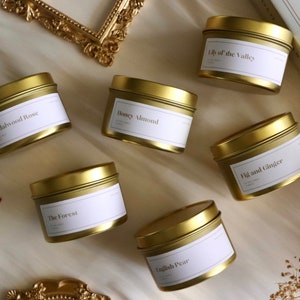 May include: Six gold metal candle tins with white labels. The labels have the names of the scents: Sandalwood Rose, The Forest, Honey Almond, English Pear, Lily of the Valley, and Fig and Ginger.