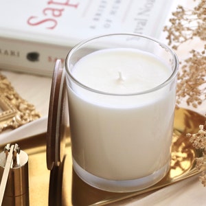 May include: A white candle in a clear glass jar with a brown lid on a gold tray. The candle is surrounded by dried flowers and a gold picture frame.