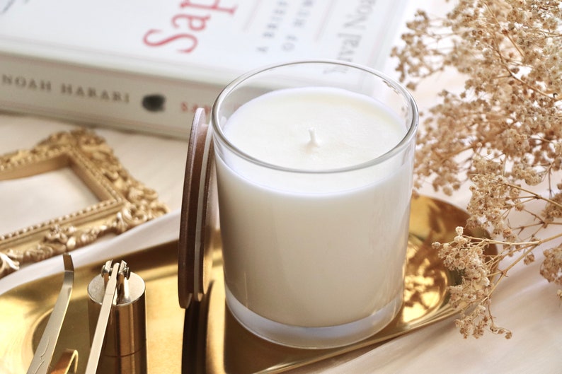 May include: A white candle in a clear glass jar with a brown lid on a gold tray. The candle is surrounded by dried flowers and a gold picture frame.