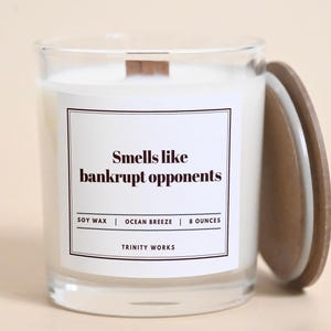 May include: A clear glass candle jar with a wooden lid. The candle is white and has a black label that reads "Smells like bankrupt opponents". The label also includes the text "Soy Wax | Ocean Breeze | 8 Ounces | Trinity Works".