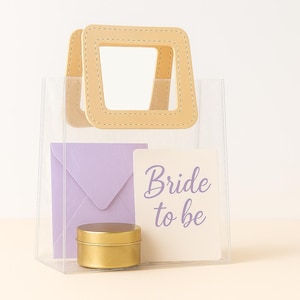 May include: A transparent tote bag with a tan handle and a lilac envelope. Inside the bag is a gold-coloured round container and a card that says "Bride to be" in a script font.