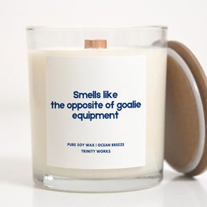 May include: A white candle in a clear glass jar with a wooden lid. The candle has a white label with the text "Smells like the opposite of goalie equipment" and "PURE SOY WAX I OCEAN BREEZE TRINITY WORKS".
