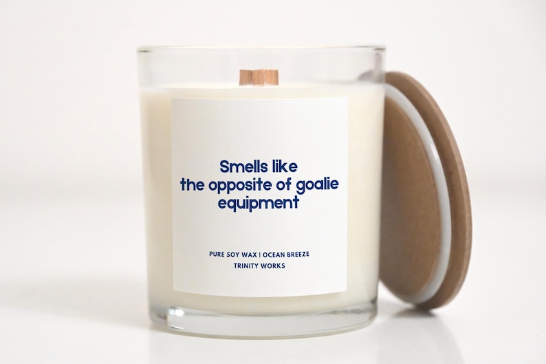 May include: A white candle in a clear glass jar with a wooden lid. The candle has a white label with the text "Smells like the opposite of goalie equipment" and "PURE SOY WAX I OCEAN BREEZE TRINITY WORKS".
