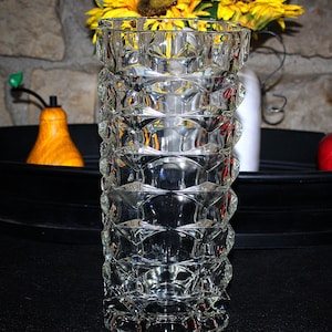 May include: A clear glass vase with a geometric pattern. The vase is tall and cylindrical with a wide mouth. The vase is made of thick glass and has a textured surface.