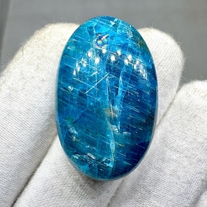 Puede incluir: Una piedra preciosa ovalada pulida en tonos azules y turquesas. La piedra tiene una calidad translúcida con patrones internos visibles y una superficie brillante. La piedra se sostiene sobre un fondo de tela blanca.