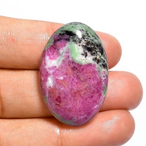 May include: Oval-shaped gemstone with a vibrant mix of colors. The stone features a prominent area of deep pink-red, contrasted by patches of green, black, and white. The polished surface reflects light, highlighting the natural patterns.