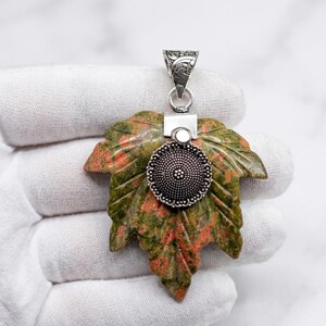 May include: A leaf-shaped pendant carved from mottled green and orange stone. The pendant features a central circular silver-toned metal embellishment with a beaded texture and a decorative silver-toned metal bail at the top for attachment.