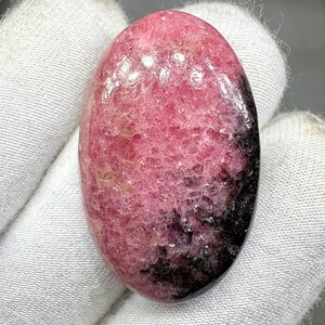 Puede incluir: Gema ovalada con un degradado de color rosa y negro. La piedra pulida tiene una superficie texturizada y se sostiene sobre un fondo de tela blanca. El tono rosa domina, pasando a negro en un extremo.