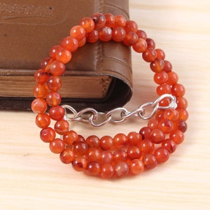 May include: A necklace made of small, round, orange-red carnelian beads. The beads are strung together with a silver-colored chain clasp. The necklace is draped over a closed, brown leather-bound book.
