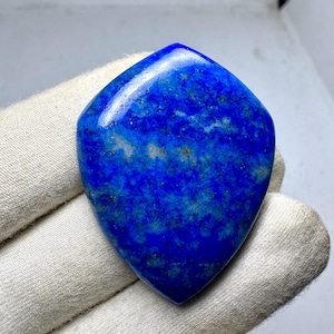 May include: A polished, shield-shaped lapis lazuli gemstone featuring a deep, vibrant blue colour with natural mottled patterns of white calcite and small flecks of gold-coloured pyrite distributed across its smooth, reflective surface.