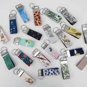 May include: A collection of colourful fabric keychains with various designs and characters, including Winnie the Pooh, Harry Potter, and Star Wars. The keychains are attached to silver key rings.