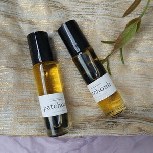 May include: Two glass roll-on bottles filled with amber liquid, each with a black cap and a white label that reads "patchouli." The bottles are positioned diagonally on a gold and white patterned fabric, with a sprig of green leaves in the background.