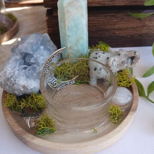 May include: A round wooden tray containing a clear glass bowl, a light blue crystal point, a cluster of blue celestite, a small stone elephant figurine with black spots, a silver crescent moon charm, and patches of bright green preserved moss.
