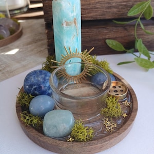 May include: A round wooden tray containing a clear glass candle holder, a tall light blue crystal point, and several polished blue stones. The tray is decorated with green moss, a gold sunburst charm, and small gold star accents.
