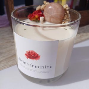 Op de afbeelding: Een heldere glazen kaars met een witte was binnenkant. De kaars is versierd met een rode roos, een lichtroze steen en gedroogde bloemen. Het label luidt "divine feminine" met een roos.