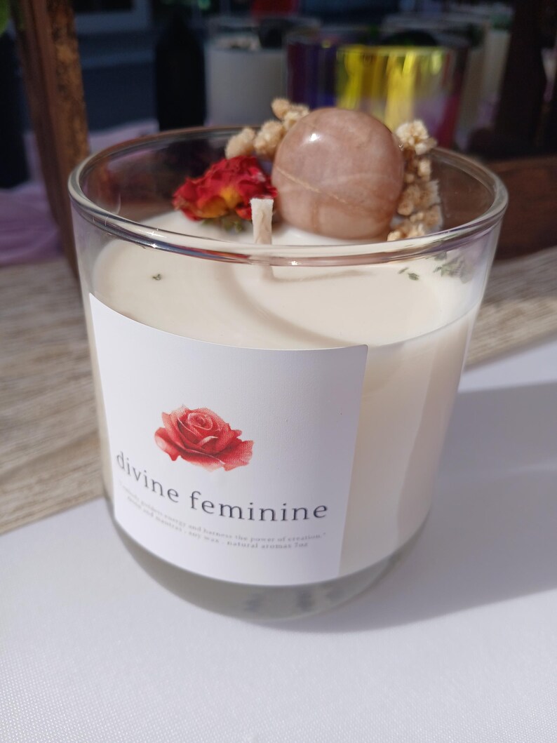 Op de afbeelding: Een heldere glazen kaars met een witte was binnenkant. De kaars is versierd met een rode roos, een lichtroze steen en gedroogde bloemen. Het label luidt "divine feminine" met een roos.