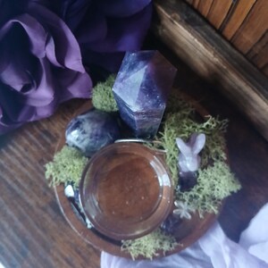 K&ouml;nnte beinhalten: Ein Holztablett mit einem Glasgef&auml;&szlig;, Amethystkristallen und einer kleinen geschnitzten Hasenfigur. Die Anordnung umfasst gr&uuml;nes Moos und lila Stoff und schafft eine mystische und dekorative Pr&auml;sentation. Die Kristalle sind tiefviolett.