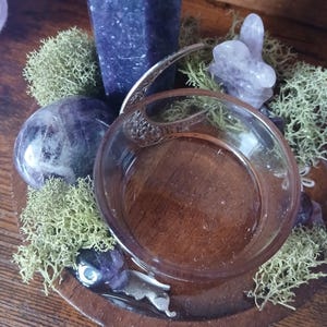 K&ouml;nnte beinhalten: Ein Holztablett mit einer Glasschale, umgeben von gr&uuml;nem Moos, Amethystkristallen und einem hohen lila Kristall. Die Schale hat einen silbernen Griff und steht auf einem Holzsockel. Die Anordnung befindet sich auf einer Holzoberfl&auml;che.