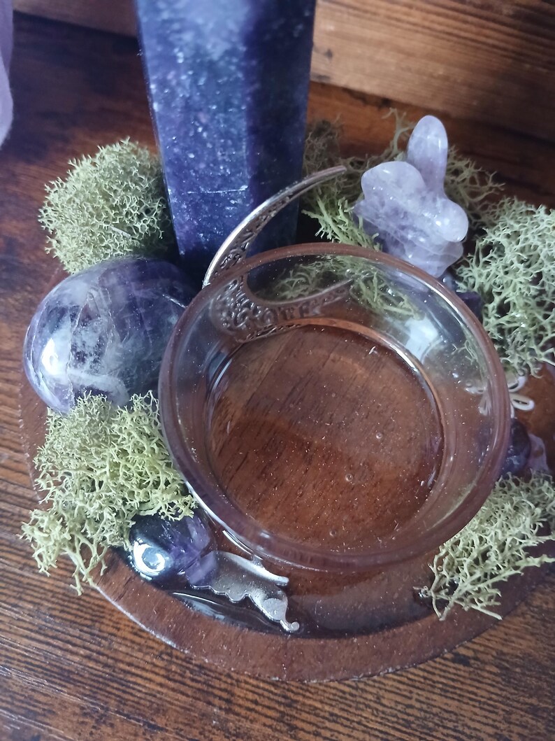 K&ouml;nnte beinhalten: Ein Holztablett mit einer Glasschale, umgeben von gr&uuml;nem Moos, Amethystkristallen und einem hohen lila Kristall. Die Schale hat einen silbernen Griff und steht auf einem Holzsockel. Die Anordnung befindet sich auf einer Holzoberfl&auml;che.