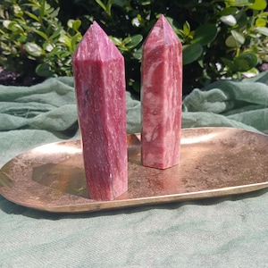 May include: Two pink and red gemstone obelisks with pointed tops sit on a gold-coloured oval tray. The obelisks have a marbled appearance, set against green foliage and a green cloth.
